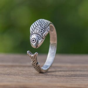 Op de afbeelding: Een zilverkleurige ring in de vorm van een vis. De kop van de vis vormt de bovenkant van de ring, de staart de onderkant. De vis heeft gedetailleerde schubben en een opvallend oog. De ring staat tegen een vage groene achtergrond.