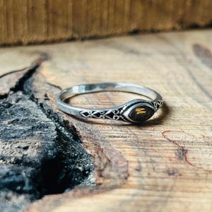 May include: A silver ring with an oval, amber gemstone. The ring band has an intricate, openwork design. The ring is set against a rustic wooden background, highlighting the jewelry's details.