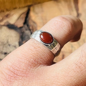 May include: Silver ring with a hammered texture and a central oval, reddish-brown stone. The ring is worn on a finger, showcasing the design and color of the gemstone. The ring's band has a textured, hammered appearance.