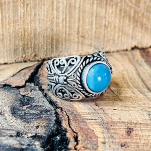 May include: A silver ring featuring an elaborate pattern and a prominent, round, bright turquoise stone. The ring has detailed scrollwork and a textured band. The ring is set against a wooden background.