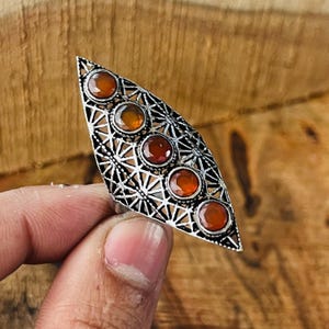 Könnte beinhalten: Ein silberfarbener Ring mit einem länglichen, blattartigen Design. Der Ring hat fünf runde, orange-rote Edelsteine, die in einem filigranen Muster gefasst sind. Der Ring wird vor einem Holzgrund gehalten.