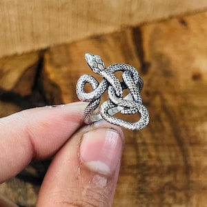 Puede incluir: Un anillo de metal plateado con forma de serpiente enroscada y una superficie texturizada que imita escamas. La cabeza de la serpiente se sitúa en la parte superior, con su cuerpo formando el aro del anillo, con detalles oxidados.