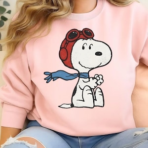May include: Pink sweatshirt featuring a cartoon Snoopy character wearing a red aviator helmet and goggles, a blue scarf, and black spots. The graphic is centered on the front of the sweatshirt.