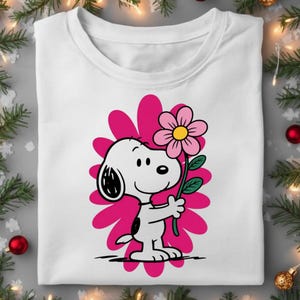 May include: White t-shirt featuring a cartoon Snoopy character holding a pink flower with a yellow center. The design includes a pink flower-shaped background. The shirt is a crew neck style.