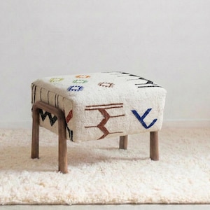 May include: A square ottoman with a cream-colored fabric cover featuring a geometric pattern in brown, blue, red, and black. The wooden legs are a dark brown color. The ottoman is placed on a cream-colored rug.
