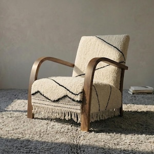 Puede incluir: Sill&oacute;n color crema con estructura y reposabrazos de madera oscura. La silla presenta una tela texturizada de color blanquecino con costuras negras y un borde con flecos. Se encuentra sobre una alfombra gris y peluda, creando un ambiente acogedor e invitador.