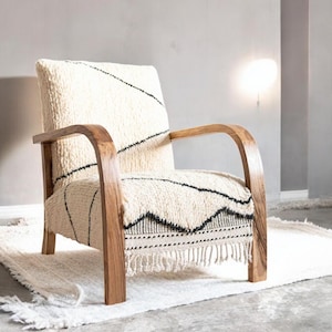 Puede incluir: Sill&oacute;n crema con estructura y reposabrazos de madera. La silla presenta una tela texturizada de color blanco roto con patrones geom&eacute;tricos negros. Se encuentra sobre una alfombra a juego con flecos. Un mueble elegante.