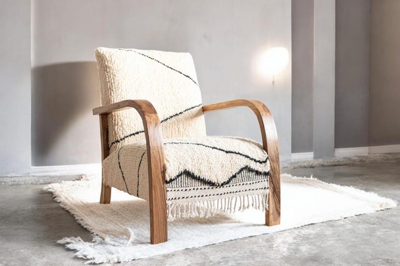 Puede incluir: Sill&oacute;n crema con estructura y reposabrazos de madera. La silla presenta una tela texturizada de color blanco roto con patrones geom&eacute;tricos negros. Se encuentra sobre una alfombra a juego con flecos. Un mueble elegante.