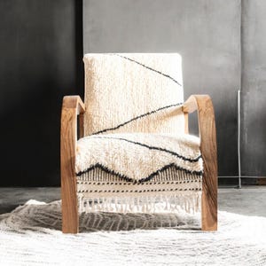 Puede incluir: Un sill&oacute;n de madera con asiento y respaldo texturizados de color crema. El asiento presenta un patr&oacute;n geom&eacute;trico en blanco y negro. La silla tiene reposabrazos y patas de madera. Debajo de la silla hay una alfombra color crema.