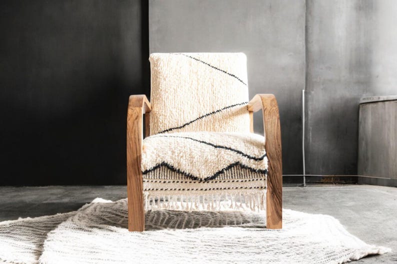 Puede incluir: Un sill&oacute;n de madera con asiento y respaldo texturizados de color crema. El asiento presenta un patr&oacute;n geom&eacute;trico en blanco y negro. La silla tiene reposabrazos y patas de madera. Debajo de la silla hay una alfombra color crema.