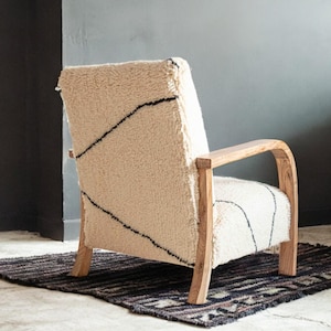 Puede incluir: Sill&oacute;n color crema con tejido texturizado y peludo y estructura de madera. La silla presenta dise&ntilde;os lineales negros en el respaldo y el asiento. Se encuentra sobre una alfombra oscura con un patr&oacute;n geom&eacute;trico, contra una pared gris oscuro.