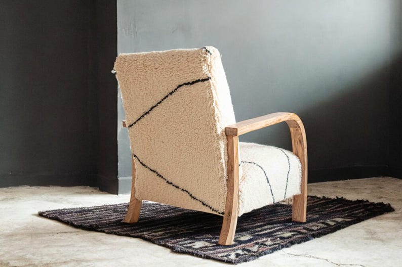 Puede incluir: Sill&oacute;n color crema con tejido texturizado y peludo y estructura de madera. La silla presenta dise&ntilde;os lineales negros en el respaldo y el asiento. Se encuentra sobre una alfombra oscura con un patr&oacute;n geom&eacute;trico, contra una pared gris oscuro.