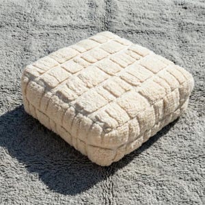 May include: A square, off-white, textured ottoman with a grid-like pattern. The ottoman is made of a soft, plush material and sits on a gray, shaggy rug. The lighting creates a shadow on the rug.