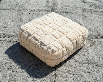May include: A square, off-white, textured ottoman with a grid-like pattern. The ottoman is made of a soft, plush material and sits on a gray, shaggy rug. The lighting creates a shadow on the rug.
