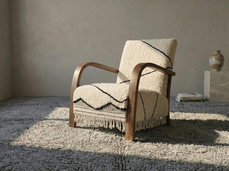 Puede incluir: Sill&oacute;n color crema con estructura y reposabrazos de madera oscura. La silla presenta una tela texturizada de color blanquecino con costuras negras y un borde con flecos. Se encuentra sobre una alfombra gris y peluda, creando un ambiente acogedor e invitador.