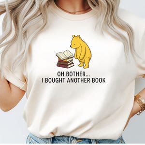 May include: A cream-colored t-shirt featuring a graphic of a yellow bear standing next to a stack of books. Below the illustration, the text reads: OH BOTHER... I BOUGHT ANOTHER BOOK. The shirt has short sleeves and a crew neckline, providing a casual look.