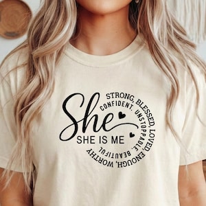 May include: A cream-colored t-shirt with the word "She" in a cursive font, encircled by words like "Strong," "Confident," and "Beautiful." The shirt's design includes two small heart shapes. The overall theme is inspirational.