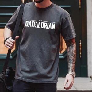 May include: A charcoal gray short-sleeve t-shirt featuring the white text THE DADALORIAN printed across the chest. The graphic includes a small silhouette of a figure standing within the letter A. The shirt has a crew neckline and a relaxed fit.