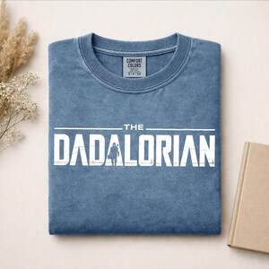 May include: A blue crewneck t-shirt with a distressed white graphic print that reads THE DADALORIAN, featuring a silhouette of a character in the center of the text. The shirt has a Comfort Colors label visible at the inner neckline.