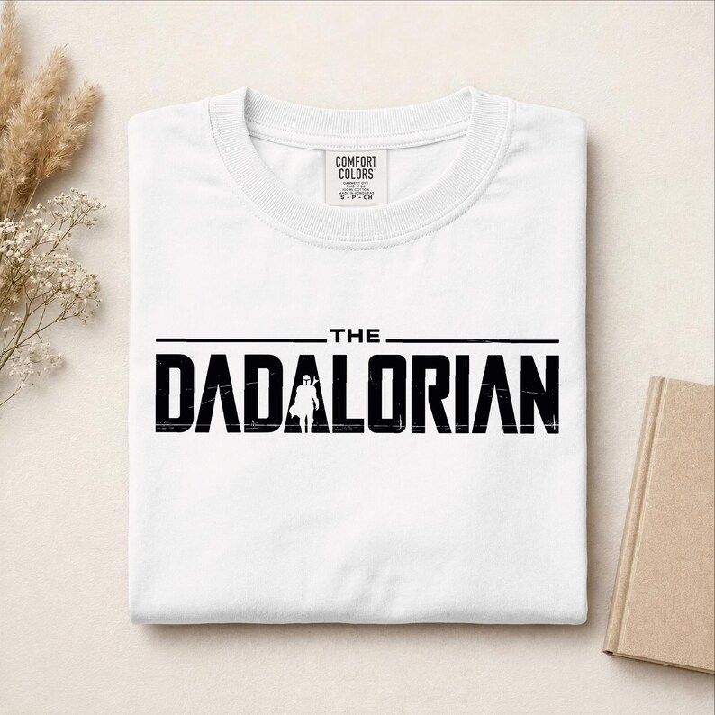 May include: A white short-sleeved t-shirt with a crew neckline and a Comfort Colors brand label at the inner collar. The front features black distressed-style text that reads THE DADALORIAN, with a silhouette of a figure replacing the letter A.