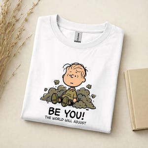 May include: A white crew neck t-shirt featuring a graphic of a cartoon character sitting in a pile of dust. Below the character, the text reads BE YOU! THE WORLD WILL ADJUST. The shirt has a Gildan brand label visible at the inner neckline.