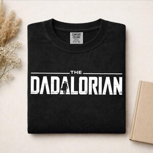 May include: A black crewneck t-shirt featuring white distressed text that reads THE DADALORIAN, with a silhouette of a helmeted figure replacing the letter A. The shirt has a Comfort Colors brand label visible inside the neckline.