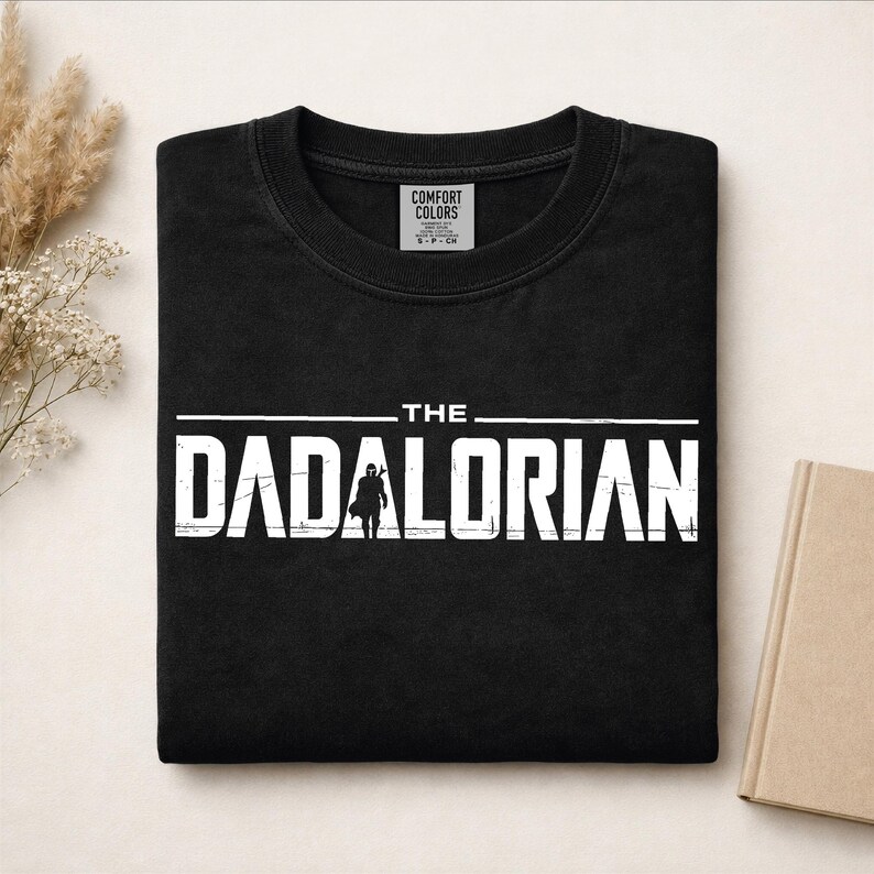May include: A black crewneck t-shirt featuring white distressed text that reads THE DADALORIAN, with a silhouette of a helmeted figure replacing the letter A. The shirt has a Comfort Colors brand label visible inside the neckline.