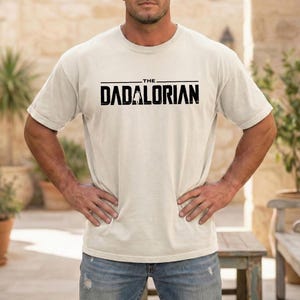 May include: A cream-colored short-sleeve t-shirt featuring the black text THE DADALORIAN printed across the chest. The graphic includes a small silhouette of a figure standing between the letters D and A. The shirt has a crew neckline.