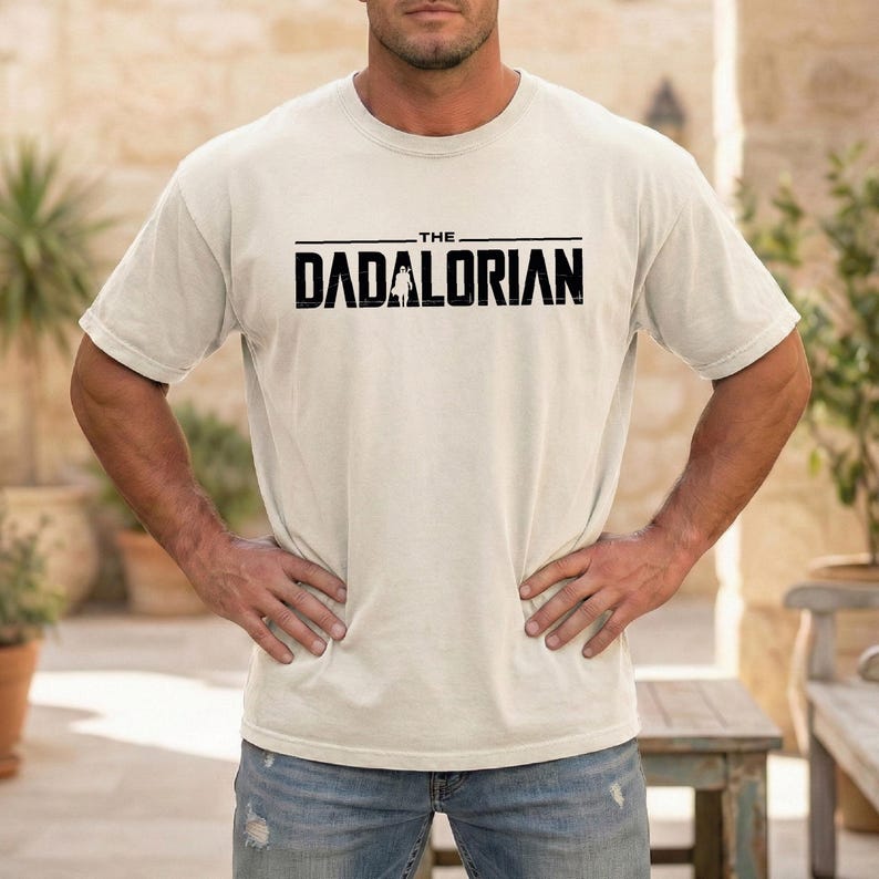 May include: A cream-colored short-sleeve t-shirt featuring the black text THE DADALORIAN printed across the chest. The graphic includes a small silhouette of a figure standing between the letters D and A. The shirt has a crew neckline.