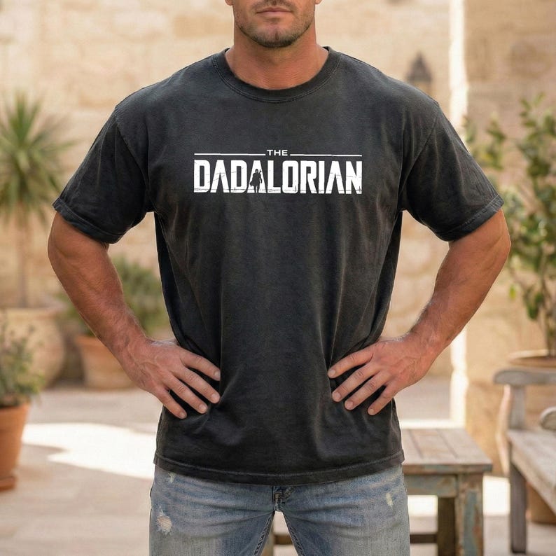 May include: A black short-sleeved crew neck t-shirt featuring the white text THE DADALORIAN printed across the chest. The graphic includes a small silhouette of a figure standing between the letters, all set against a solid dark background.