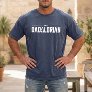 May include: A blue short-sleeved crew neck t-shirt featuring the white text THE DADALORIAN printed across the chest. The graphic includes a small silhouette of a figure standing in the center of the word, set against a solid blue fabric background.