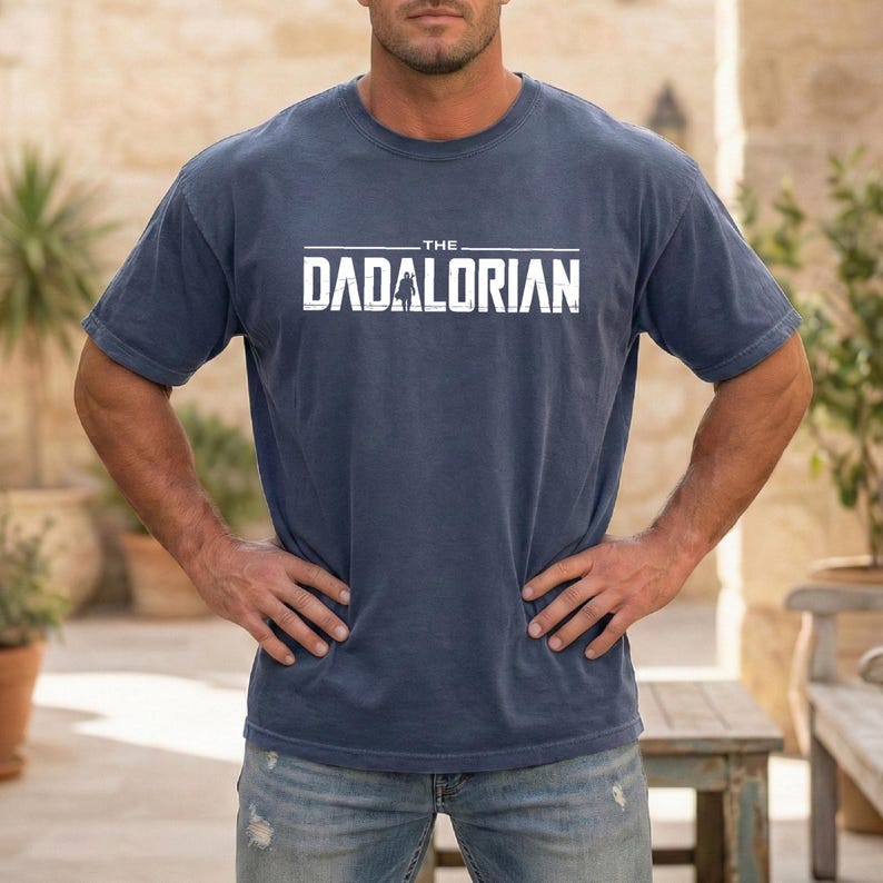 May include: A blue short-sleeved crew neck t-shirt featuring the white text THE DADALORIAN printed across the chest. The graphic includes a small silhouette of a figure standing in the center of the word, set against a solid blue fabric background.