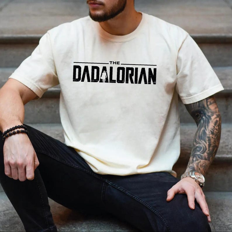May include: A cream-colored short-sleeve t-shirt featuring the black text THE DADALORIAN printed across the chest. The letter A in the word DADALORIAN is replaced by a silhouette of a figure wearing a helmet and cape, styled in a bold font.