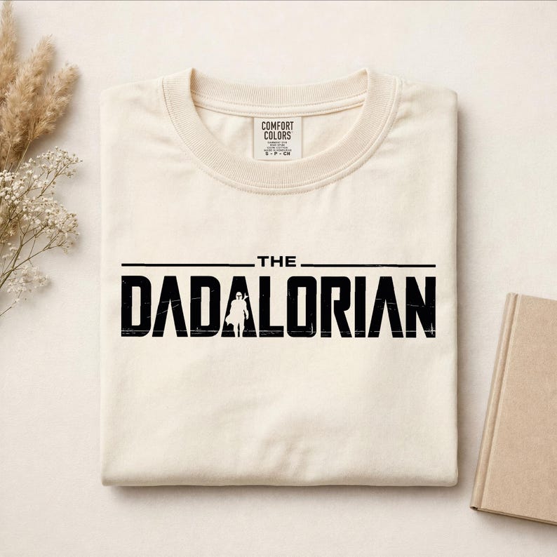 May include: A cream-colored t-shirt featuring the black text THE DADALORIAN printed across the chest. The letter A in the word DADALORIAN is replaced by a silhouette of a helmeted figure. The shirt has a Comfort Colors brand label at the neck.