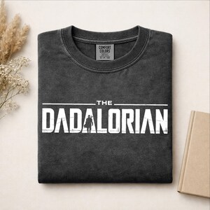 May include: A charcoal gray crewneck t-shirt with a distressed white graphic print that reads THE DADALORIAN, featuring a silhouette of a character in the center of the text. The shirt has a ribbed collar and a Comfort Colors brand neck label.