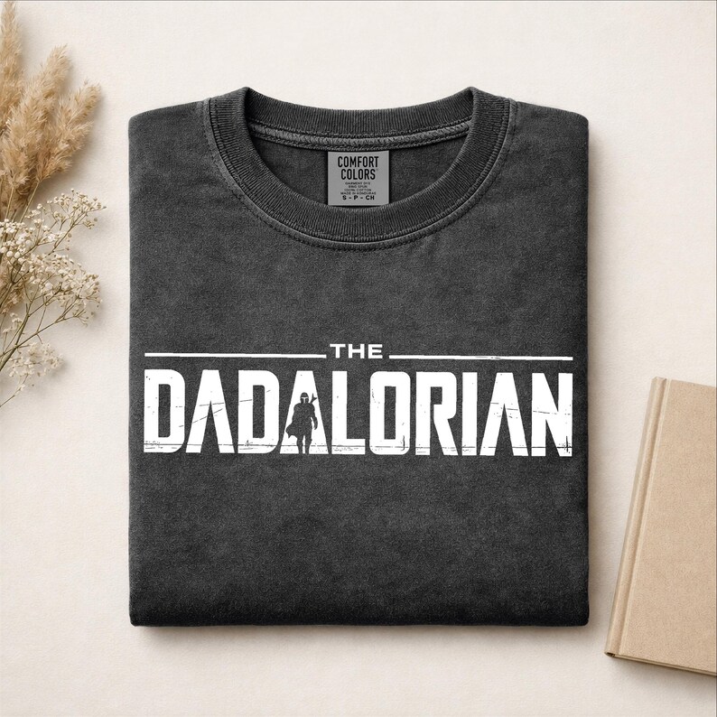 May include: A charcoal gray crewneck t-shirt with a distressed white graphic print that reads THE DADALORIAN, featuring a silhouette of a character in the center of the text. The shirt has a ribbed collar and a Comfort Colors brand neck label.