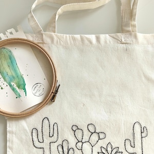 Può includere: Borsa tote color crema con un design di cactus ricamato in nero. Un acquerello di un cactus in un telaio da ricamo in legno è appoggiato sulla borsa. La borsa ha lunghe cinghie.
