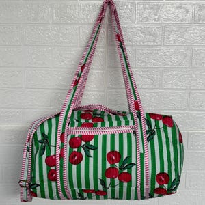 Puede incluir: Bolso tote a rayas verdes y blancas con un estampado de cerezas. El bolso tiene cierre de cremallera y ribete rosa. Las asas son de rayas rosas y blancas.