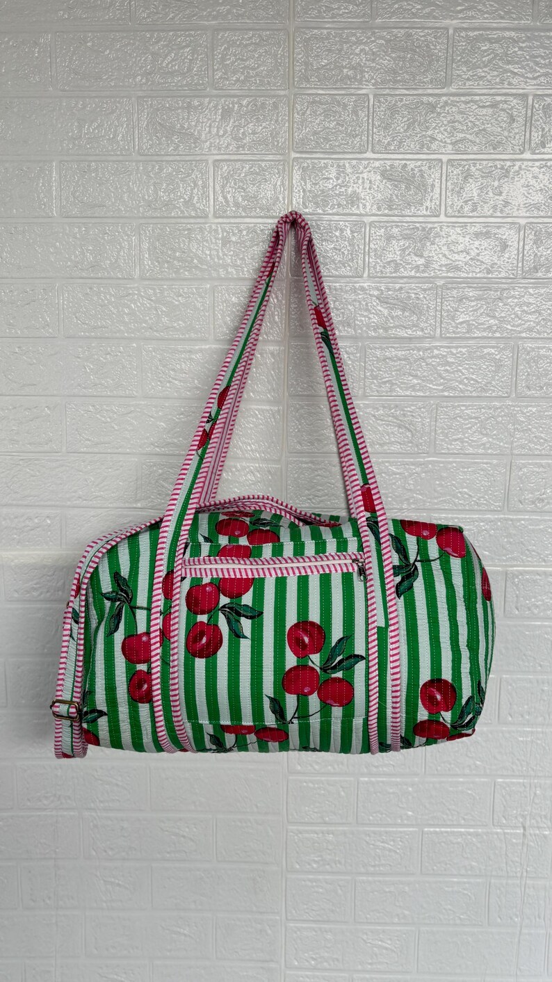 Puede incluir: Bolso tote a rayas verdes y blancas con un estampado de cerezas. El bolso tiene cierre de cremallera y ribete rosa. Las asas son de rayas rosas y blancas.