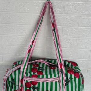Puede incluir: Un bolso tote a rayas verdes y blancas con un estampado de cerezas. El bolso tiene un ribete y correas rosas. El bolso est&aacute; colgado contra una pared de ladrillos blancos.