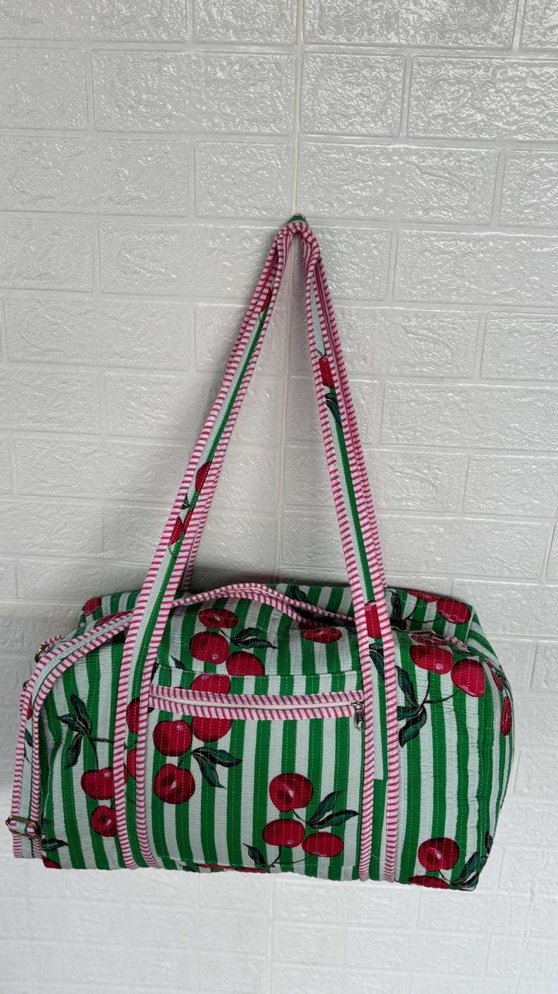 Puede incluir: Un bolso tote a rayas verdes y blancas con un estampado de cerezas. El bolso tiene un ribete y correas rosas. El bolso est&aacute; colgado contra una pared de ladrillos blancos.