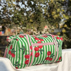 Puede incluir: Bolsa de lona a rayas verdes y blancas con estampado de cerezas. La bolsa tiene un bolsillo con cremallera y ribete rosa. La bolsa est&aacute; hecha de una tela suave y tiene dos asas.