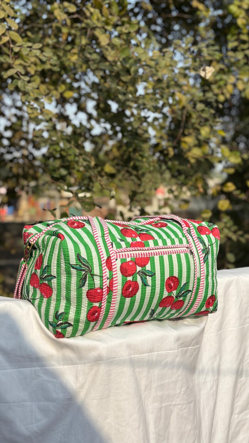 Puede incluir: Bolsa de lona a rayas verdes y blancas con estampado de cerezas. La bolsa tiene un bolsillo con cremallera y ribete rosa. La bolsa est&aacute; hecha de una tela suave y tiene dos asas.