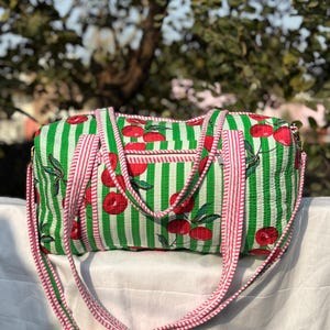 Puede incluir: Una bolsa de lona a rayas verdes y blancas con un estampado de cerezas. La bolsa tiene un bolsillo con cremallera y una larga correa para el hombro a rayas rosas y blancas. La bolsa est&aacute; hecha de tela acolchada.