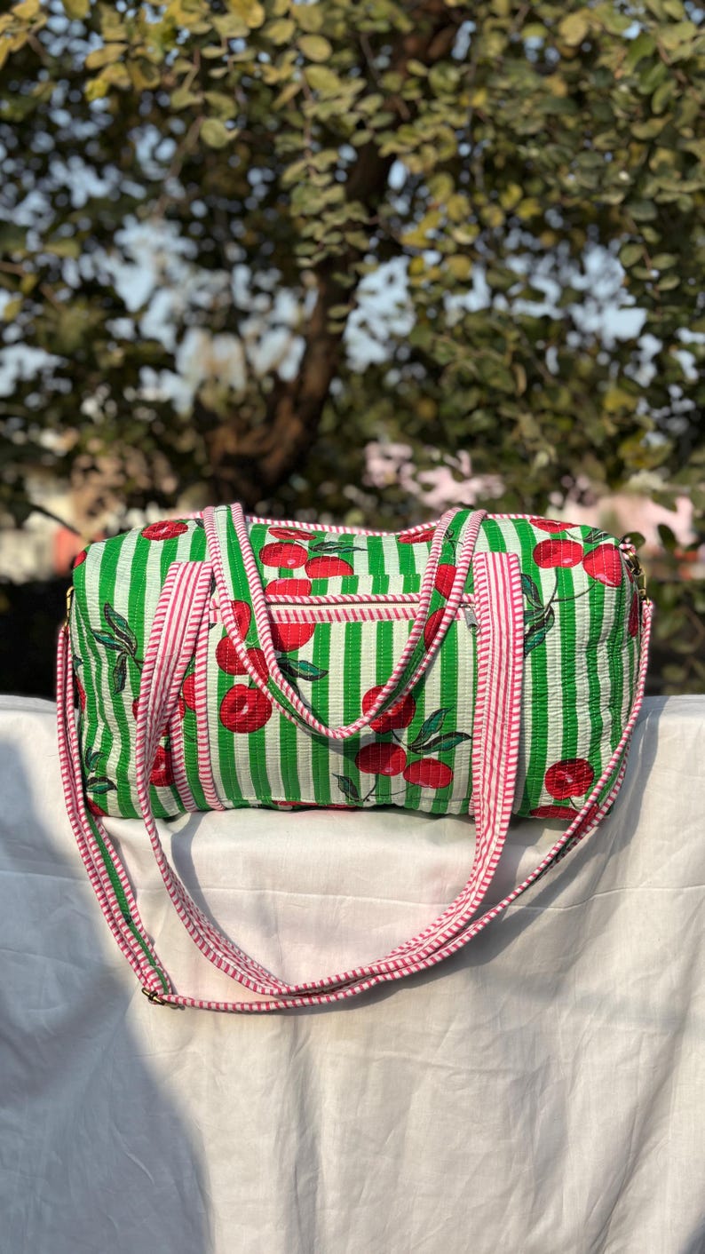 Puede incluir: Una bolsa de lona a rayas verdes y blancas con un estampado de cerezas. La bolsa tiene un bolsillo con cremallera y una larga correa para el hombro a rayas rosas y blancas. La bolsa est&aacute; hecha de tela acolchada.