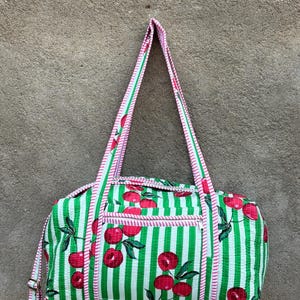 Puede incluir: Una bolsa de lona a rayas verdes y blancas con un estampado de cerezas. La bolsa tiene forma rectangular con un bolsillo con cremallera y dos asas. Las asas y los ribetes son rosas.