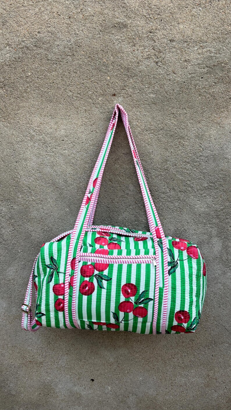 Puede incluir: Una bolsa de lona a rayas verdes y blancas con un estampado de cerezas. La bolsa tiene forma rectangular con un bolsillo con cremallera y dos asas. Las asas y los ribetes son rosas.