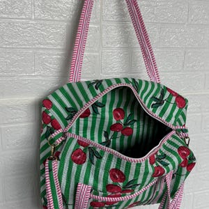Puede incluir: Bolso tote a rayas verdes y blancas con un estampado de cerezas. El bolso tiene una correa y ribete a rayas rosas y blancas. El bolso est&aacute; abierto, revelando un interior espacioso. Cuelga contra una pared de ladrillos blancos.
