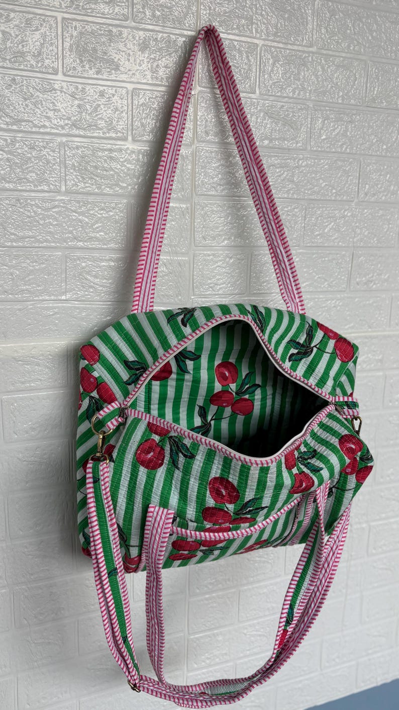 Puede incluir: Bolso tote a rayas verdes y blancas con un estampado de cerezas. El bolso tiene una correa y ribete a rayas rosas y blancas. El bolso est&aacute; abierto, revelando un interior espacioso. Cuelga contra una pared de ladrillos blancos.