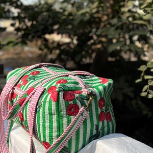 Puede incluir: Una bolsa de lona a rayas verdes y blancas con un estampado de cerezas. La bolsa tiene una correa a rayas rosas y blancas y una cremallera dorada. La bolsa est&aacute; sobre una superficie blanca.